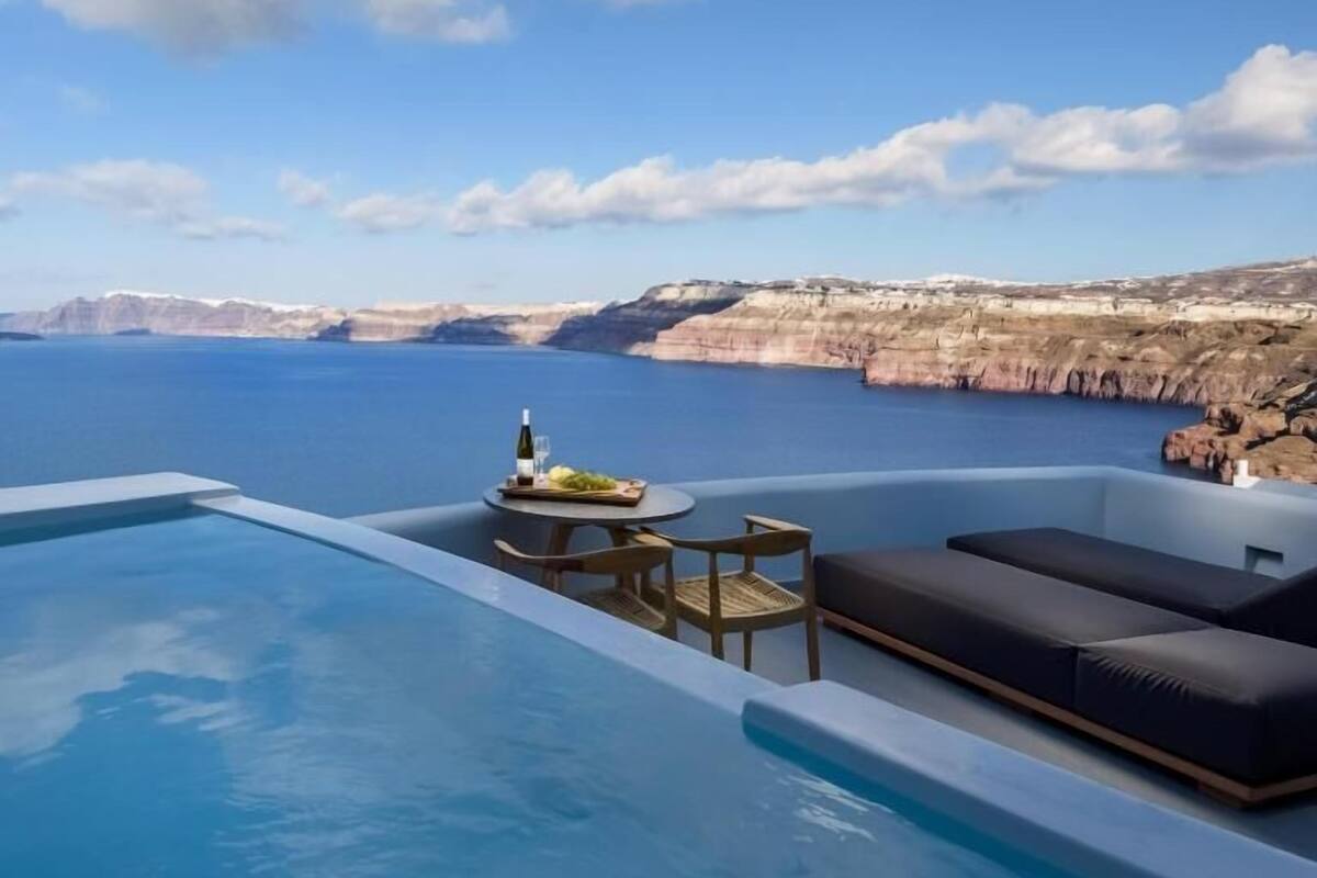 Una piscina con vistas al mar y a los acantilados, una mesa con una botella y fruta, y un área de descanso.