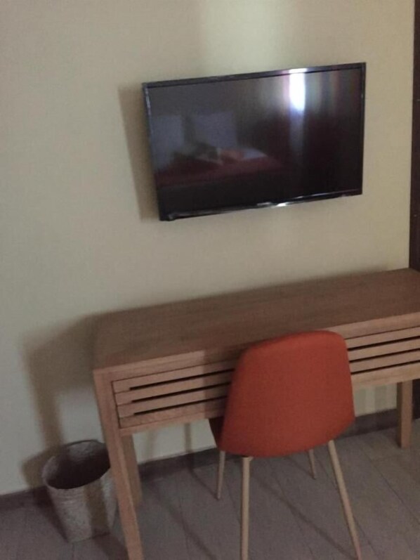 Una habitación con un escritorio de madera, una silla roja, un televisor montado en la pared y un cubo de basura.