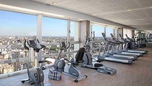 Une salle de sport équipée de tapis de course, d’elliptiques et de vélos d’exercice, avec de grandes fenêtres offrant une vue sur la ville.
