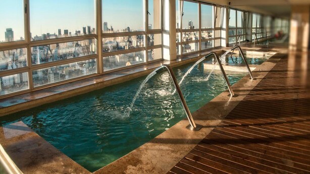 Une piscine intérieure offrant une vue dégagée sur la ville grâce à de grandes fenêtres, ainsi qu’une terrasse en bois et des échelles de piscine en métal.