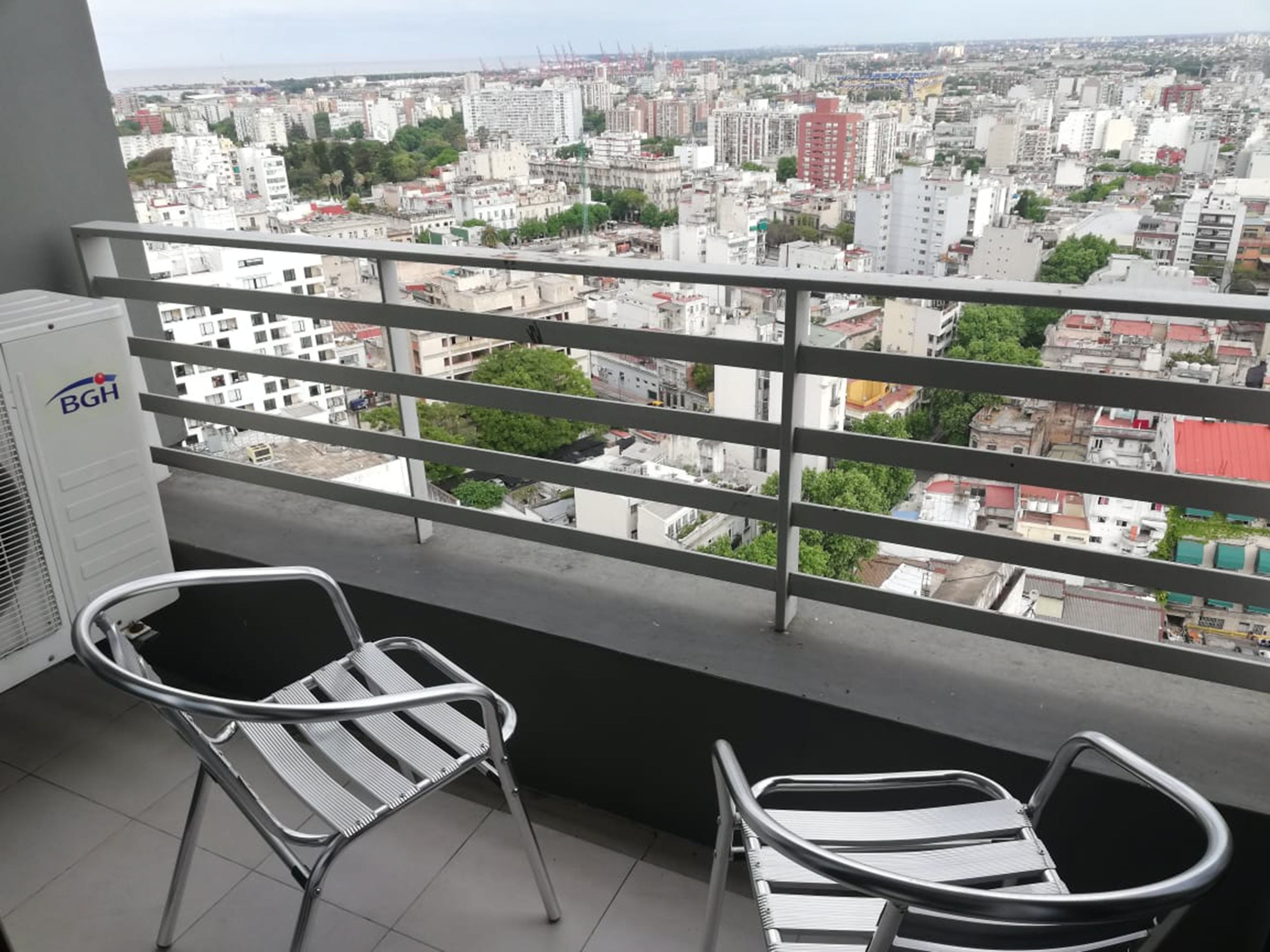 Un balcón con barandillas metálicas, dos sillas metálicas y vistas a un paisaje urbano con edificios y zonas verdes.