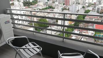 Un balcón con barandillas metálicas, dos sillas metálicas y vistas a un paisaje urbano con edificios y zonas verdes.