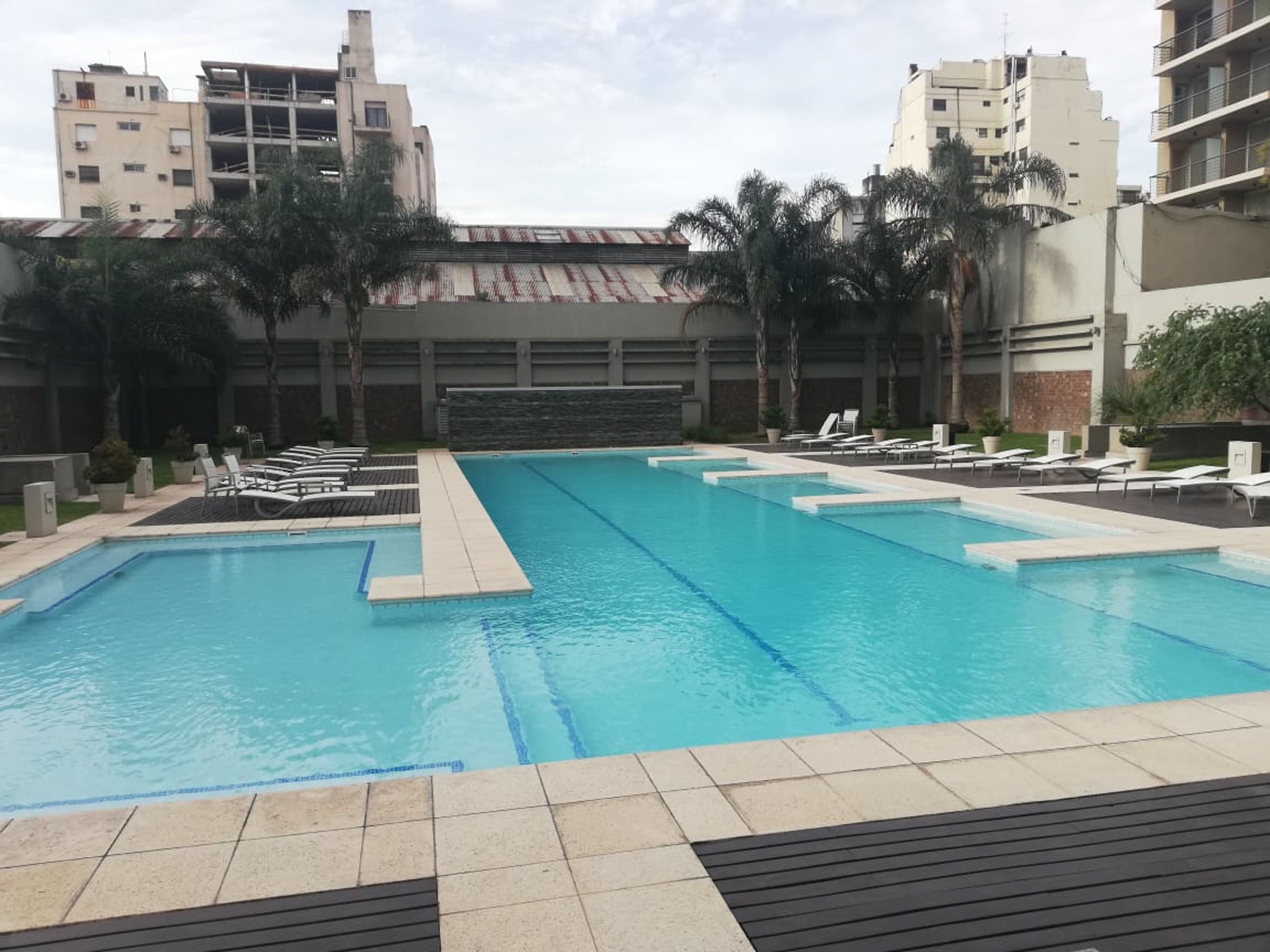 Una piscina con varias pistas de nado, rodeada de sillones de descanso y palmeras, con edificios al fondo.