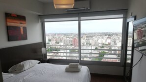 Une chambre d’hôtel dotée d’une grande fenêtre donnant sur la ville, d’un lit bien fait avec du linge de maison blanc et d’un luminaire moderne.