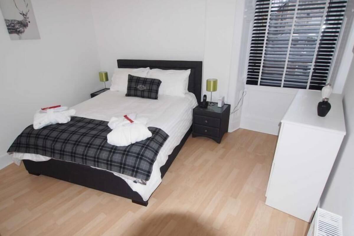 A neatly made bed with a plaid bedspread and white towels, a bedside table with a lamp, and a radiator in a room with wooden flooring.