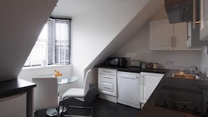 A modern kitchen with white cabinetry, a black countertop, and a small dining area with a glass table and white chair. - Thistle Apartments (Aberdeen)