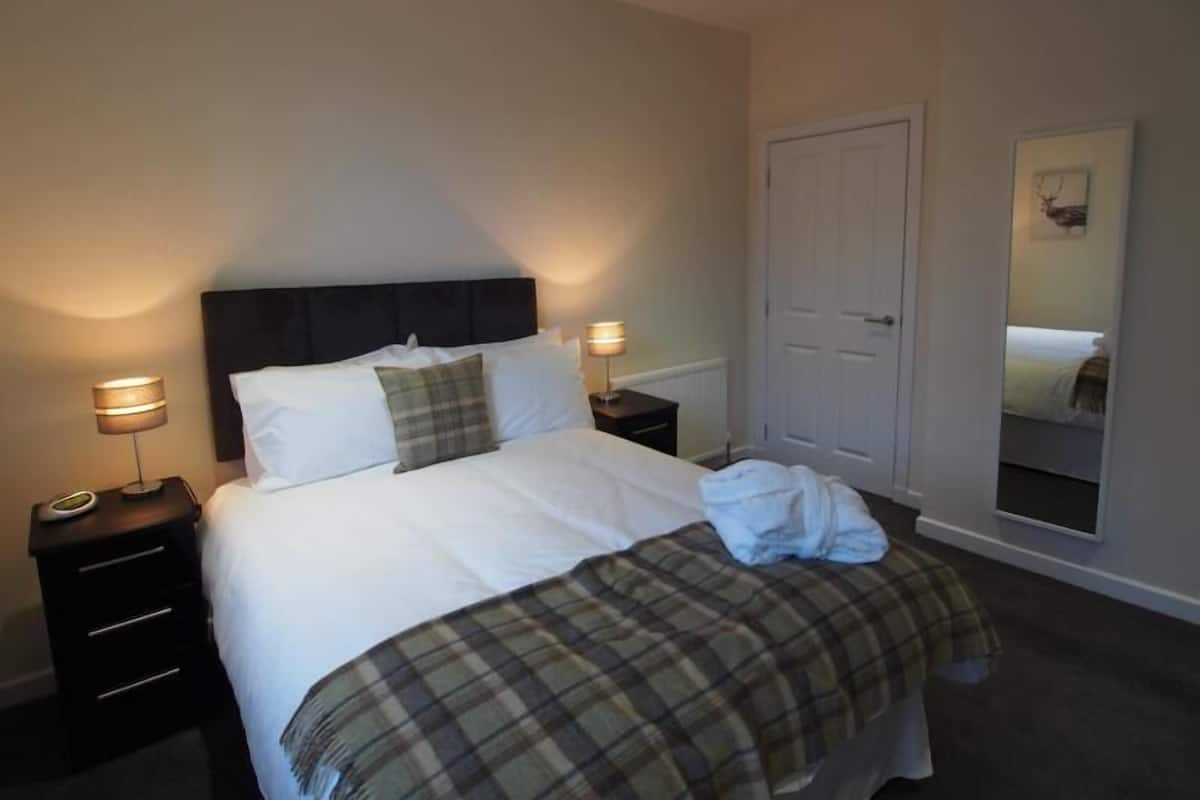 A neatly made bed with a plaid bedspread, two bedside lamps, a dark headboard, and a white door.