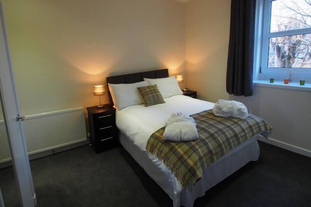 A neatly made bed with a plaid blanket, two bedside lamps, and a window with curtains.