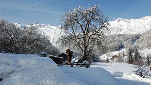 Paisagem coberta de neve com uma cabana de madeira, uma árvore e montanhas ao fundo.