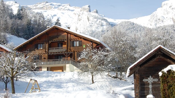 Um chalé de madeira com varanda, rodeado por árvores e montanhas cobertas de neve.