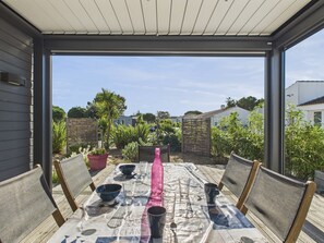 Eine Terrasse im Freien mit einem Esstisch, umgeben von Stühlen, und Blick auf einen Garten sowie Gebäude im Hintergrund.