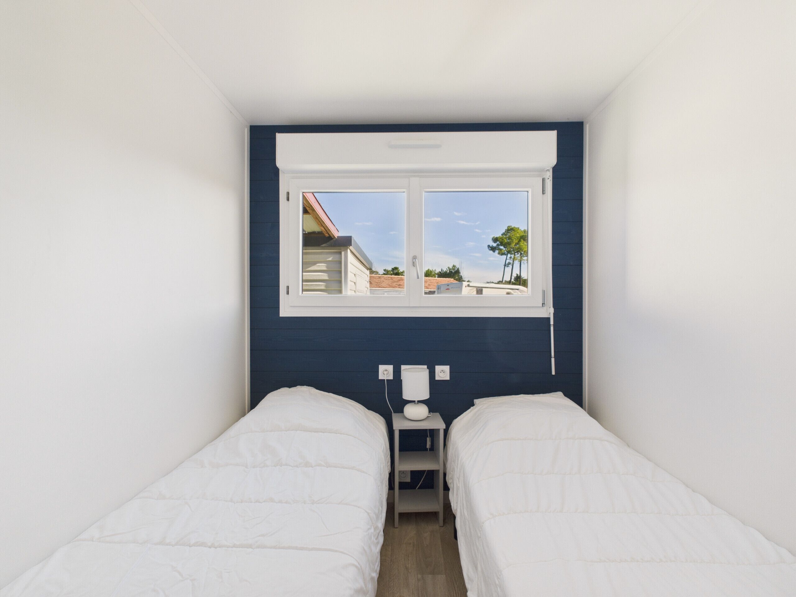 Un dormitorio con dos camas individuales, una ventana con vistas a edificios y una mesita de noche con lámpara.
