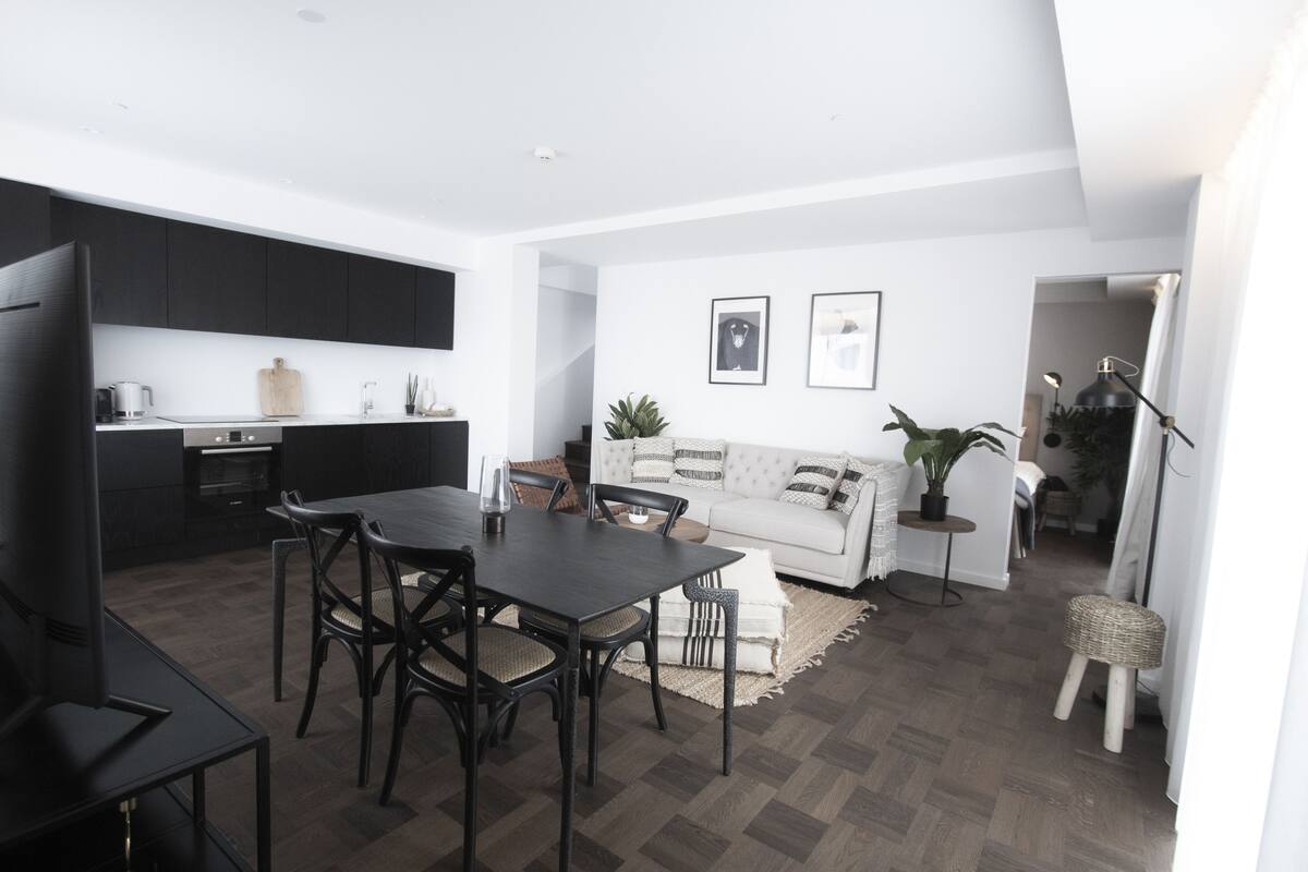 Una cocina moderna con armarios oscuros, una mesa de comedor con sillas y un sofá blanco en una sala de estar luminosa.