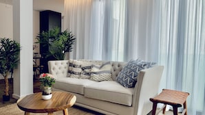 A living room with a beige sofa, patterned cushions, a wooden coffee table, a small wooden stool, and two potted plants. - Terreno Suites (Palma de Mallorca)