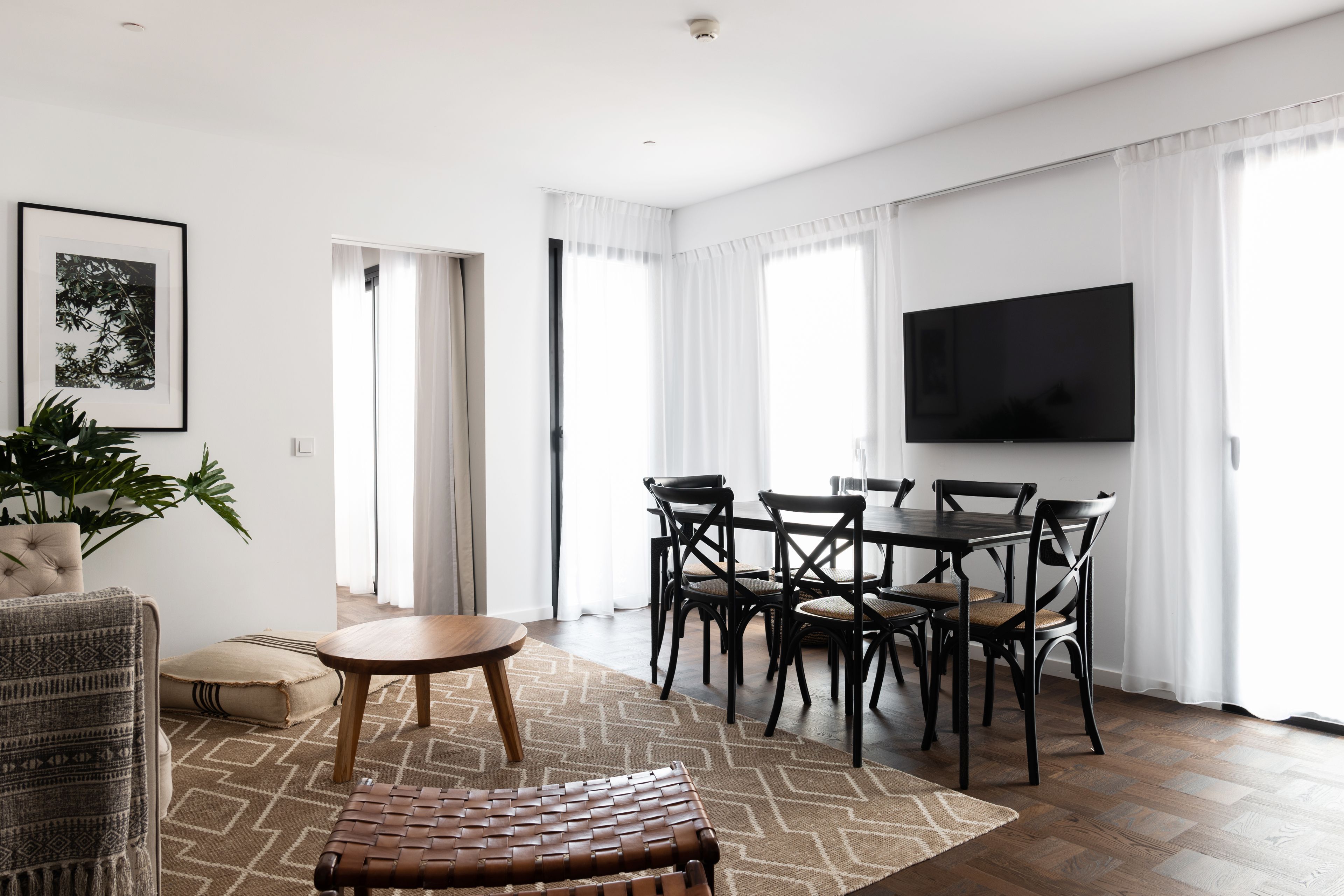 A modern living room with a dining area, a flat-screen TV, and a large window with sheer curtains.