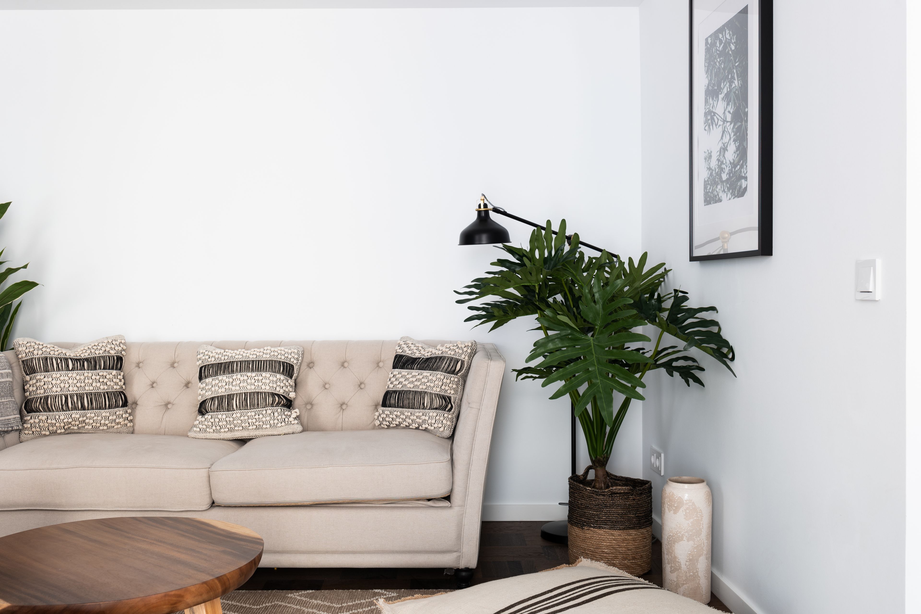 Ein Wohnzimmer mit einem beigefarbenen Sofa, einem runden Holztisch, einem Blumentopf und einem gerahmten Bild an der Wand.