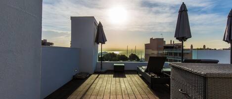 A rooftop terrace with wooden flooring, a seating area with a table, and two umbrellas.