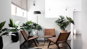 A modern indoor space with a round table and two chairs, surrounded by large potted plants, against a white wall with a black floor lamp and a window with blinds. - Terreno Suites (Palma de Mallorca)