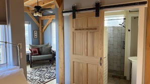 Une pièce avec une porte en bois, un ventilateur de plafond, un canapé avec des coussins, un tapis à motifs et une vue sur une salle de bain équipée d’une douche et d’un lavabo.