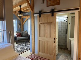 Una habitación con puerta de madera, ventilador de techo, sofá con cojines, alfombra estampada y vista a un baño con ducha y lavamanos.
