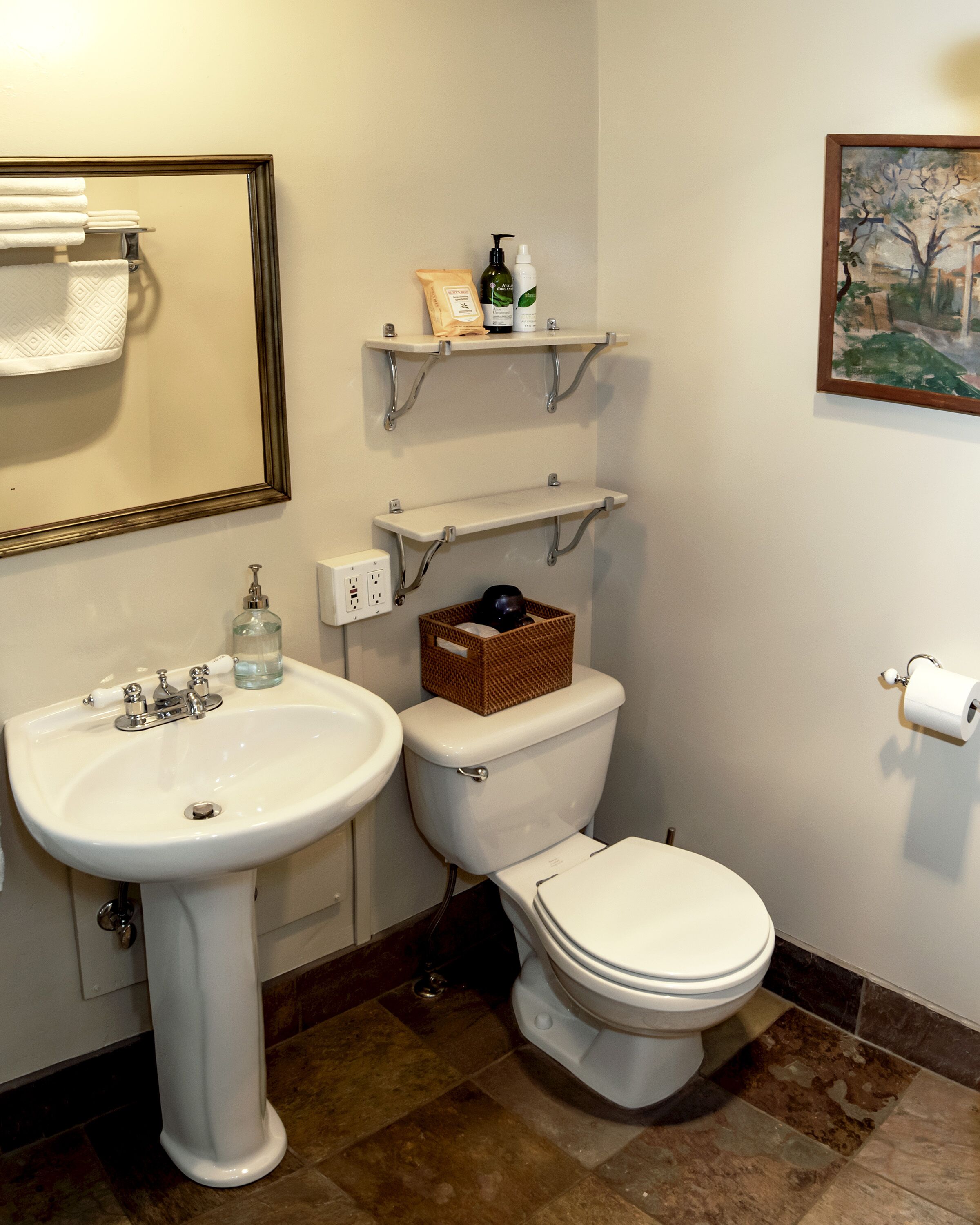 Un baño con lavamanos pedestal, inodoro y repisas con artículos de aseo.