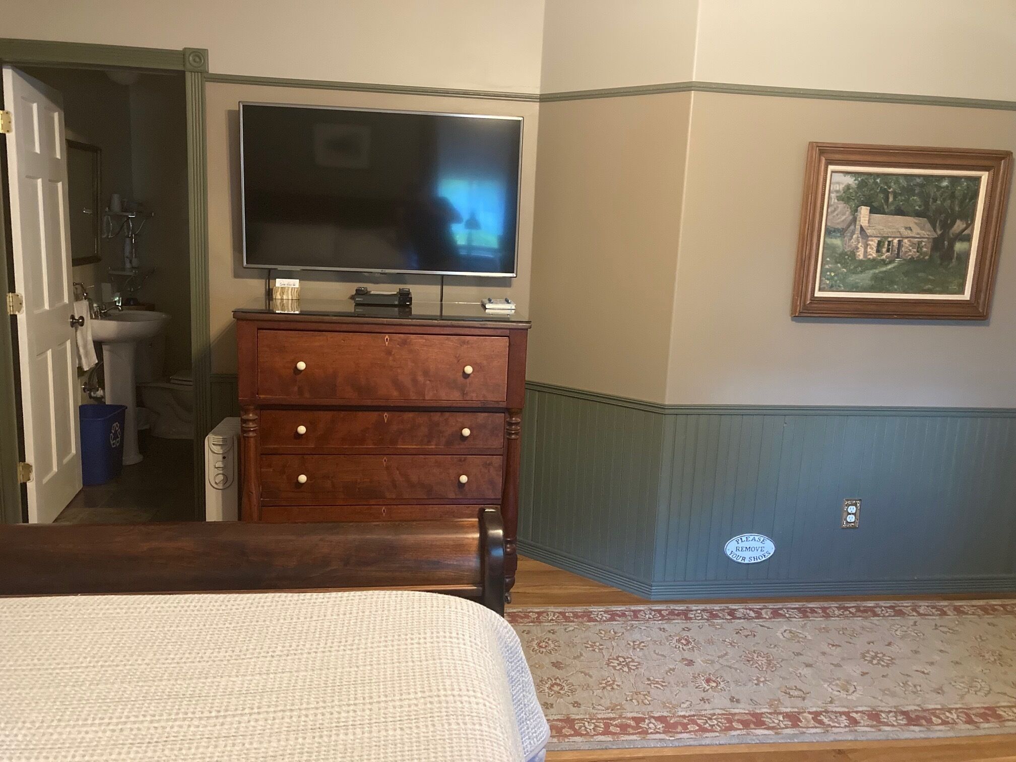 A bedroom with a bed, a dresser with a flat-screen TV, a bathroom with a sink and toilet, and a painting on the wall.