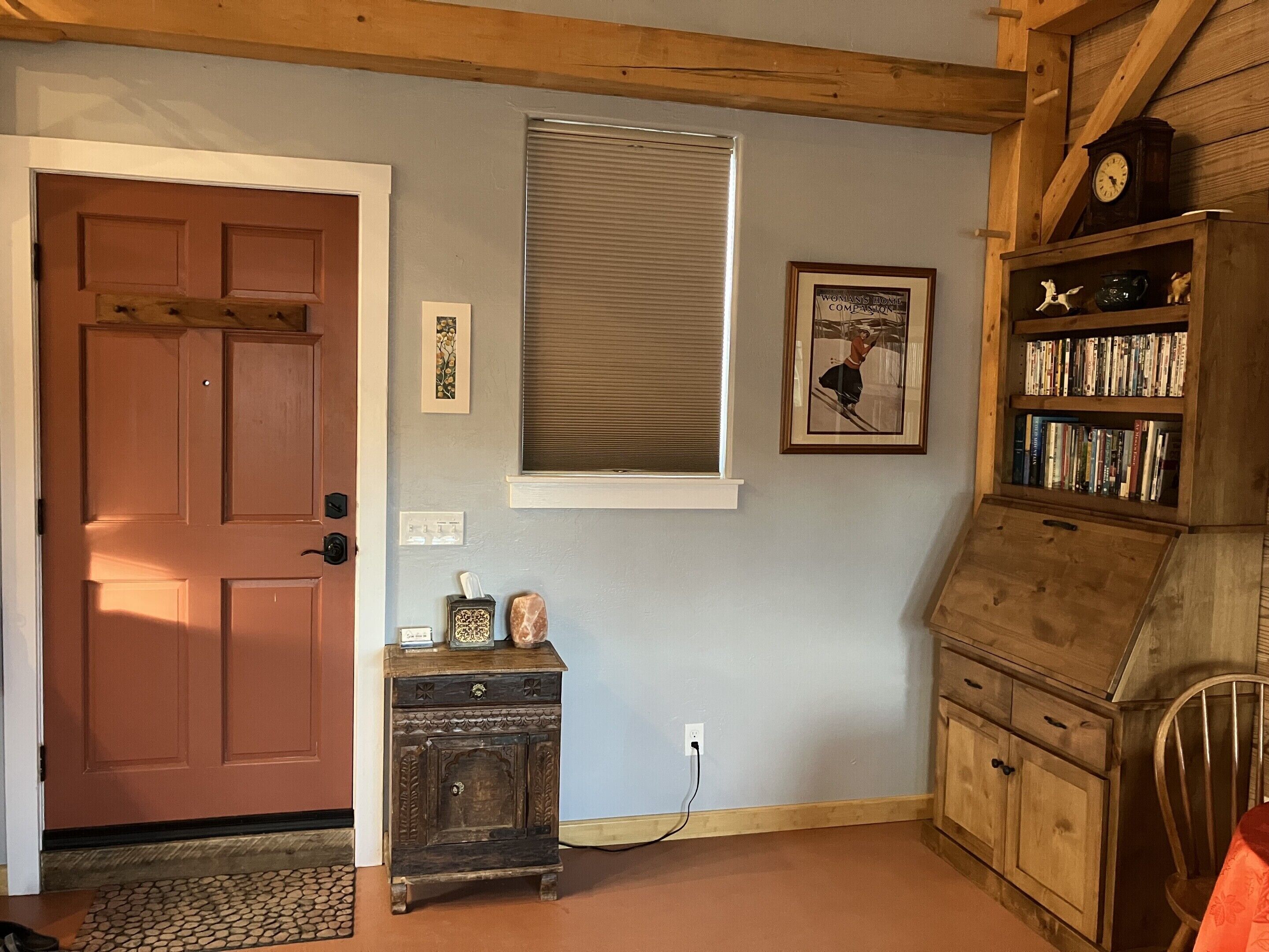 A room with a wooden door, a small wooden cabinet with a clock, a window with blinds, a framed picture on the wall, and a bookshelf filled with books.