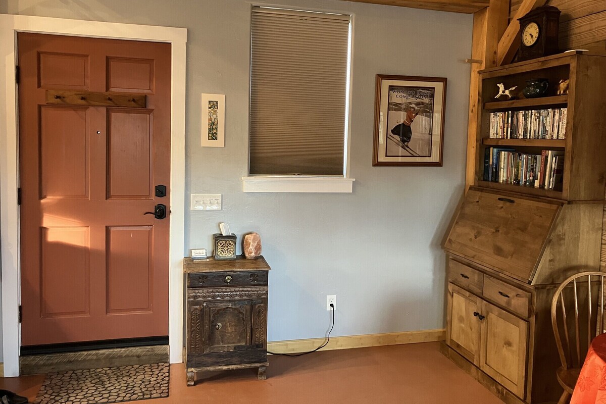 A room with a wooden door, a small wooden cabinet with a clock, a window with blinds, a framed picture on the wall, and a bookshelf filled with books.