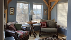 Une pièce chaleureuse avec un plafond en pente, des poutres en bois, une grande fenêtre, un canapé, un fauteuil et une petite table avec une lampe.