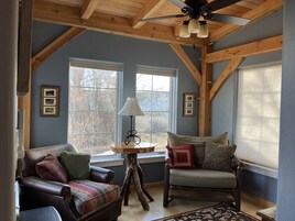 A cozy room with a sloped ceiling, wooden beams, a large window, a sofa, a chair, and a small table with a lamp.