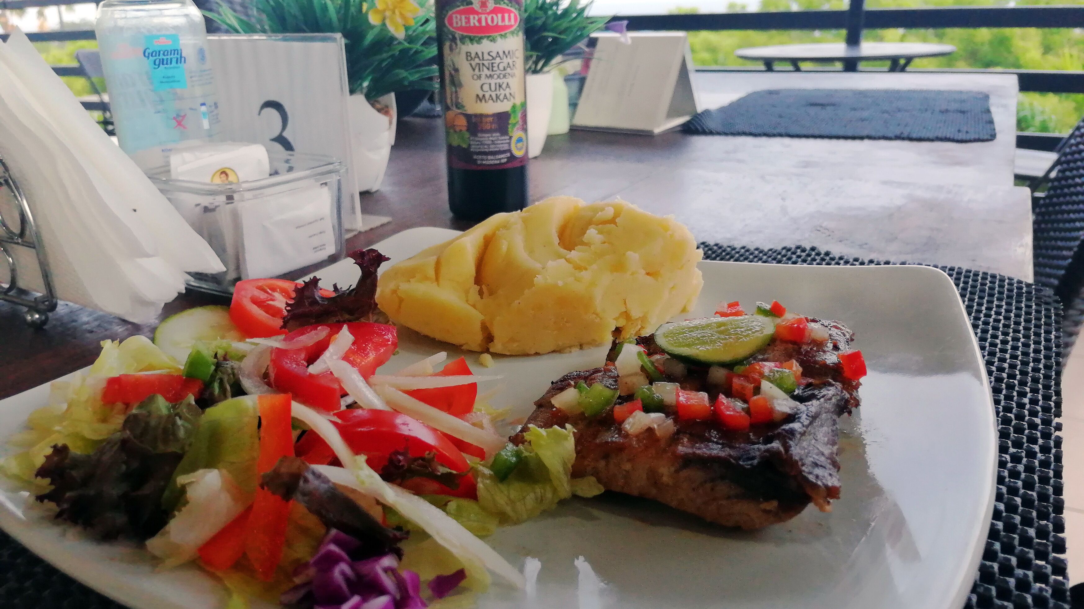 Prato com purê de batata, salada e um prato de carne, acompanhado de uma garrafa de vinagre balsâmico.