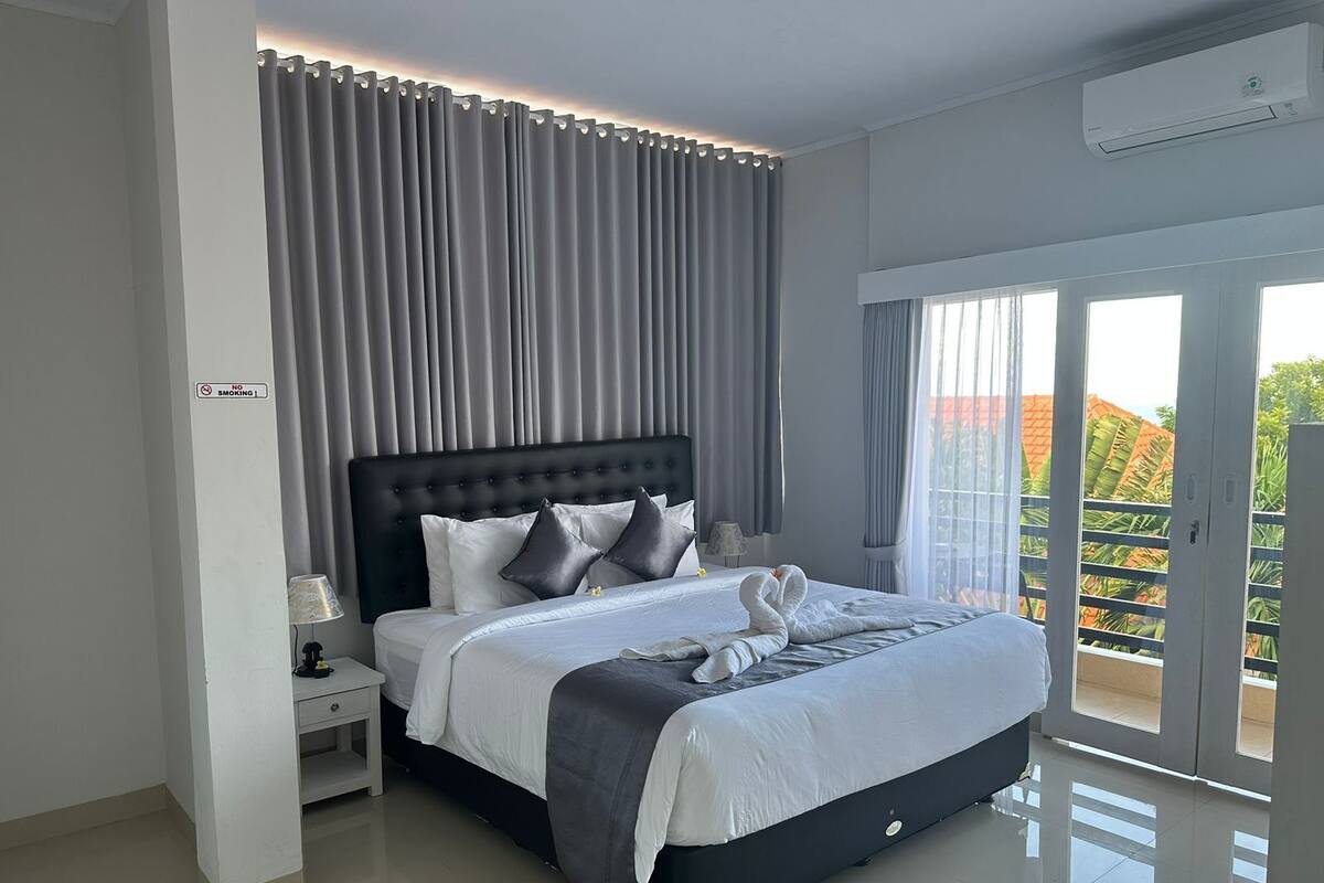 A modern hotel bedroom with a large bed, grey curtains, a no-smoking sign, a nightstand, and a view of the outdoors.