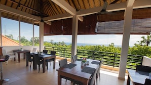 A covered outdoor dining area with tables and chairs, large windows, and a view of greenery. - Ocean Villa Dive Resort - Tulamben (Tulamben)