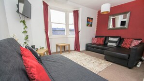 Ein modernes Wohnzimmer mit einem grauen Sofa, roten Kissen, einem kleinen Holztisch und einem großen Fenster mit roten Vorhängen.