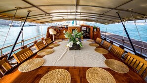 A dining area on a boat with a table set for six, surrounded by wooden chairs, a central floral arrangement, and a view of the water and cityscape. - Venezia Boat San Marco (Venice)