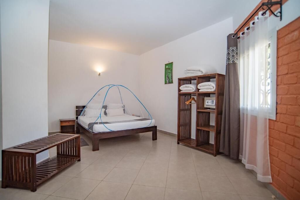 Un dormitorio con cama, mesita de noche, banco, armario y ventana con cortinas.