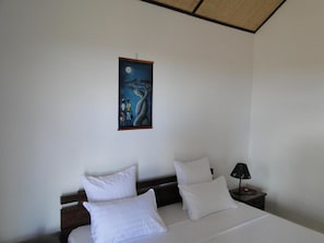 Quarto com cama, duas almofadas brancas, mesa de cabeceira com abajur e um quadro de uma árvore e duas figuras na parede.