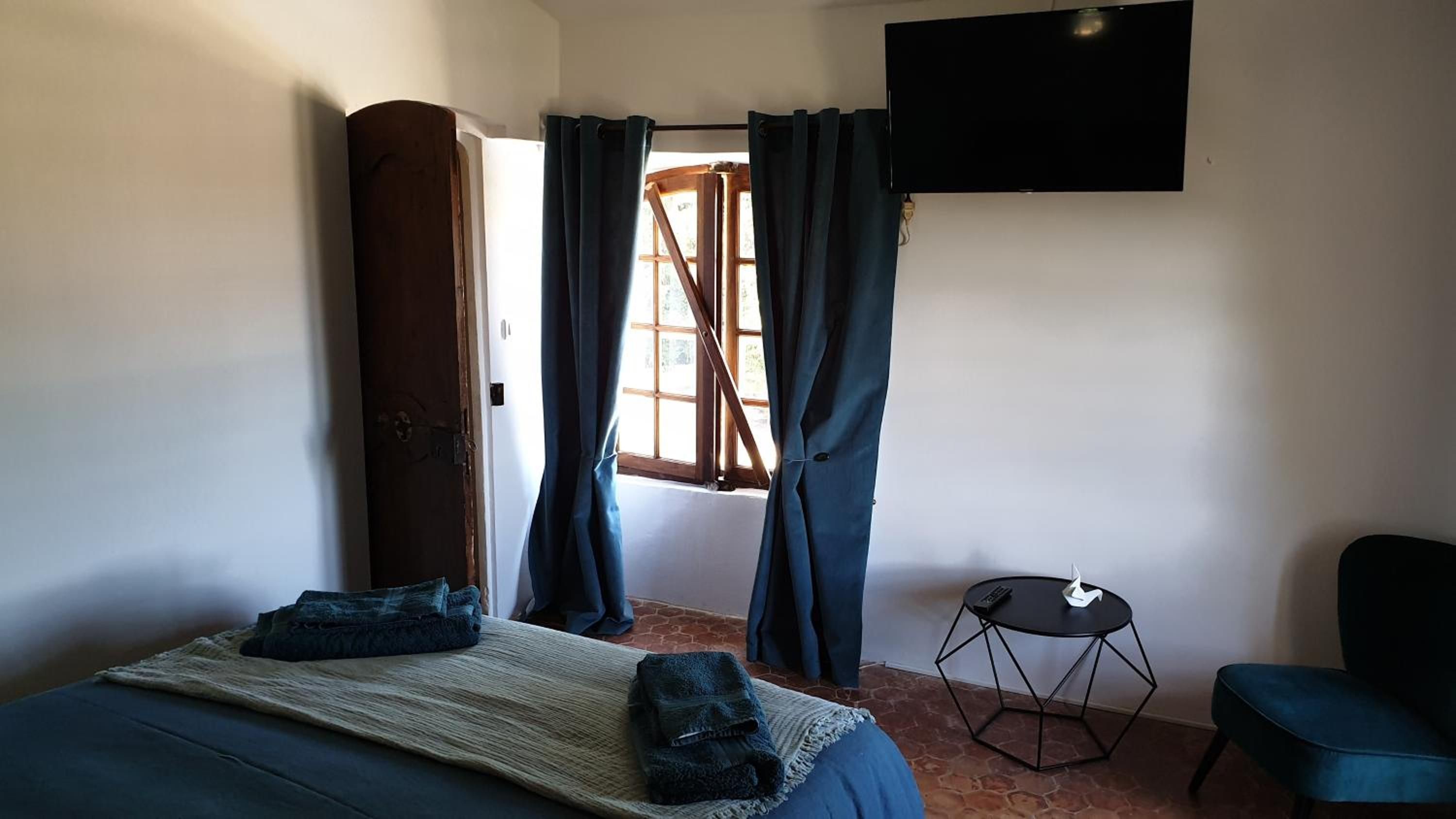 Un dormitorio con cama, una mesita redonda, una silla, un televisor y una ventana con cortinas.