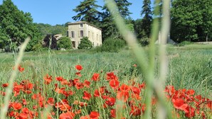 A field of red poppies in front of a stone building with a tiled roof, surrounded by trees. - Bastide de l'Adrech (Manosque)