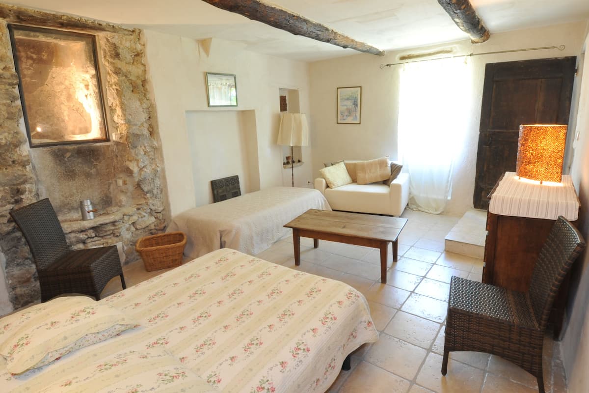 Una habitación acogedora con cama, sofá, sillón y mesa. Se aprecia una pared de piedra y una viga de madera en el techo.