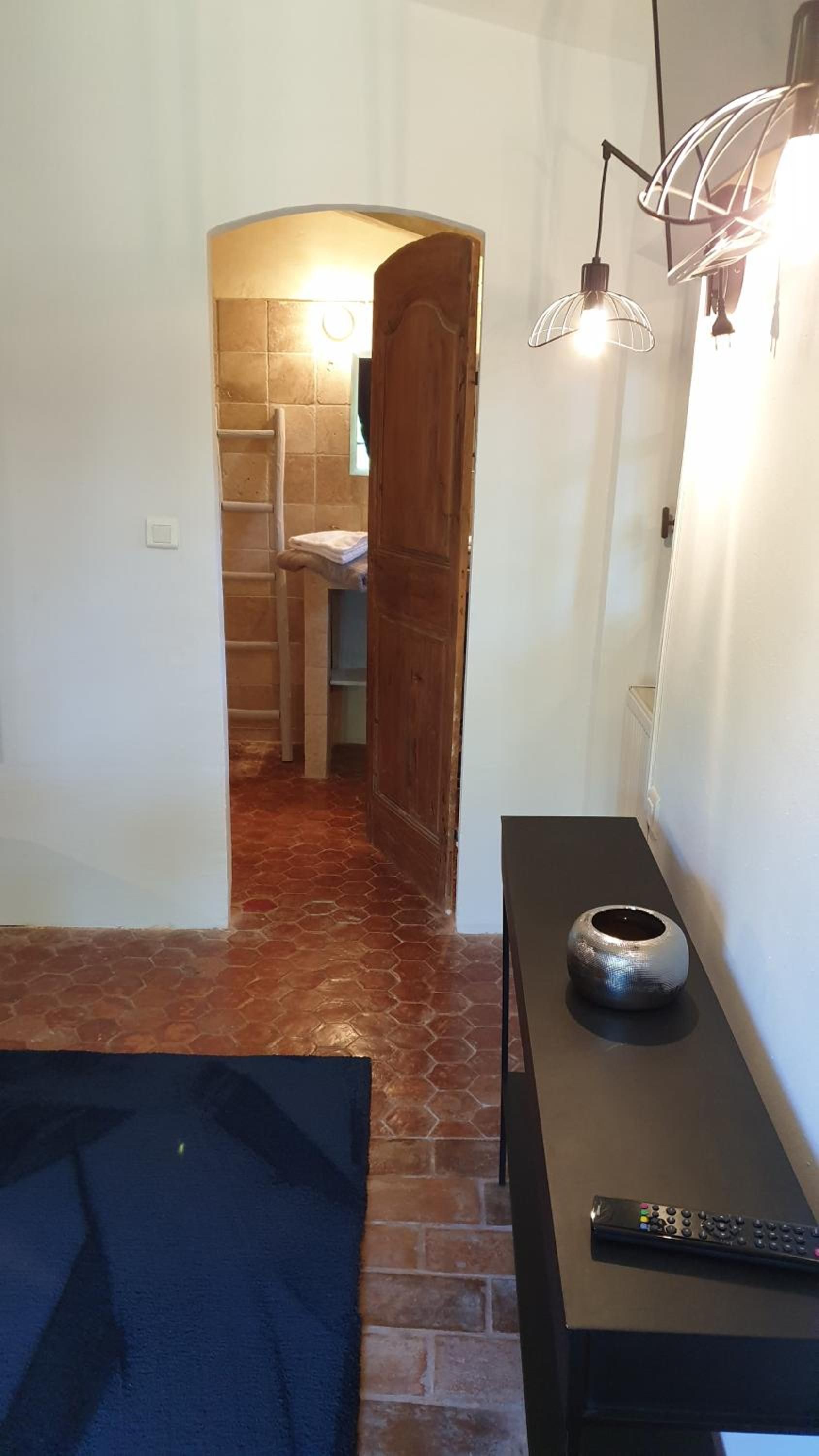 A hallway with a tiled floor, a wooden door, a small table with a remote control, and a wall-mounted light fixture.