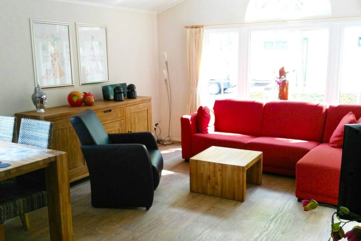 A living room with a red sofa, a wooden coffee table, a black armchair, a dining table with chairs, and a cabinet with kitchen appliances.