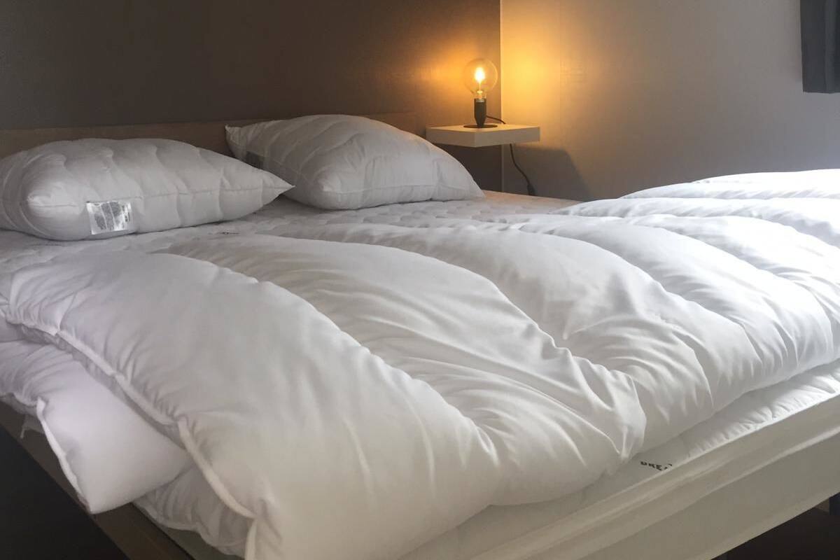 A neatly made bed with white bedding and pillows, a bedside lamp, and a plain wall.