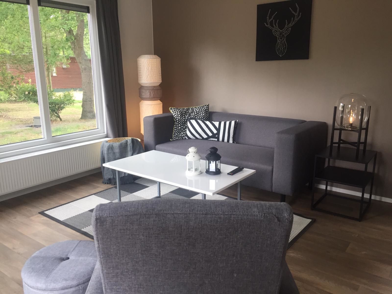 Ett modernt vardagsrum med en grå soffa, ett vitt soffbord och en grå fåtölj. Det finns en lampa, en lykta och en dekorativ väggdekoration.