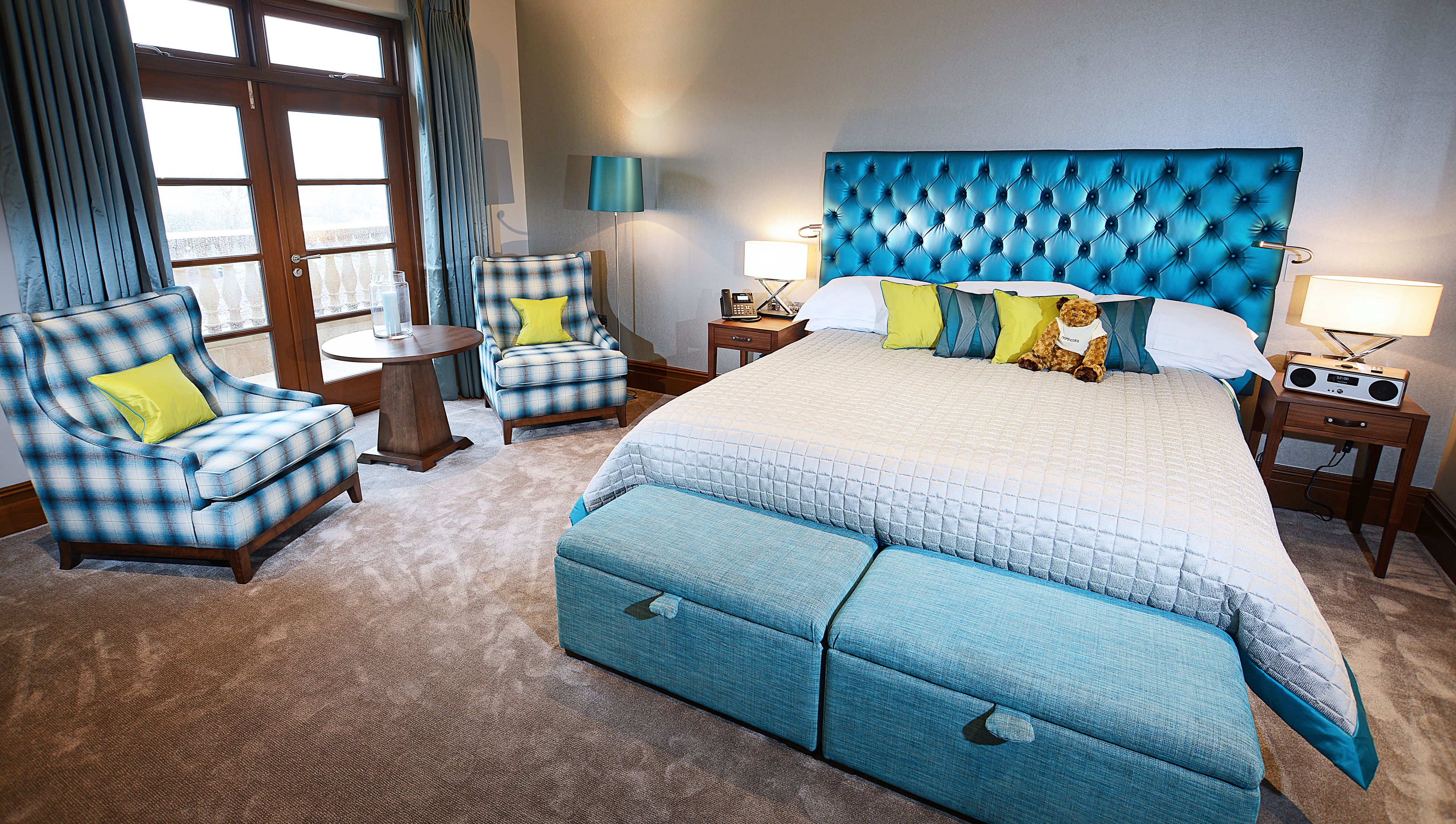 A hotel room with a large bed, a blue upholstered armchair, a matching ottoman, a small round table, and a nightstand with a lamp and radio.