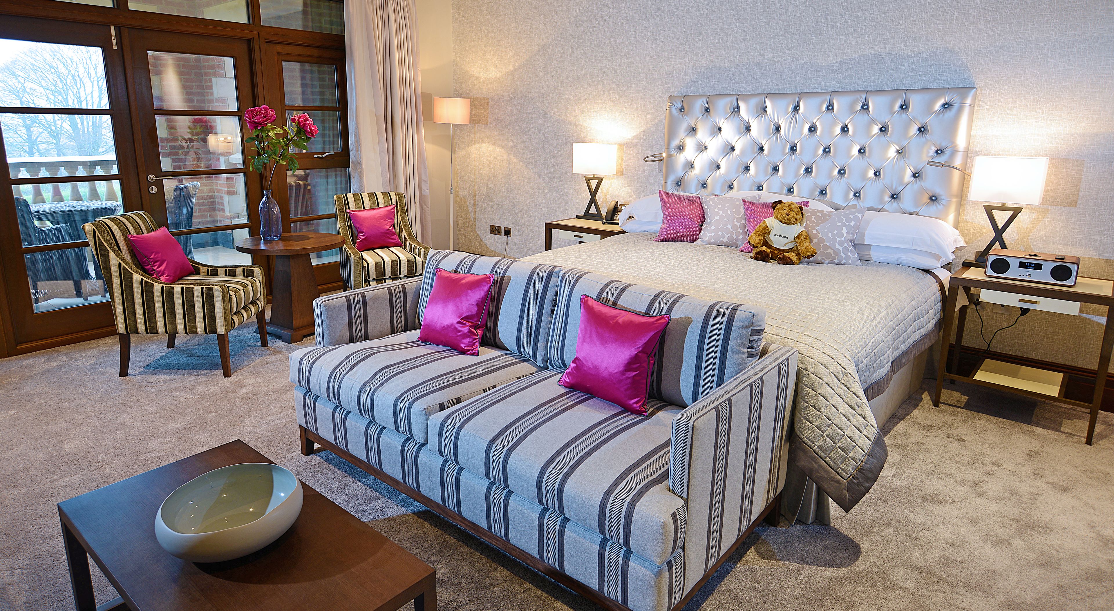 A hotel room with a bed, a sofa, a chair, a bedside table, and a lamp.