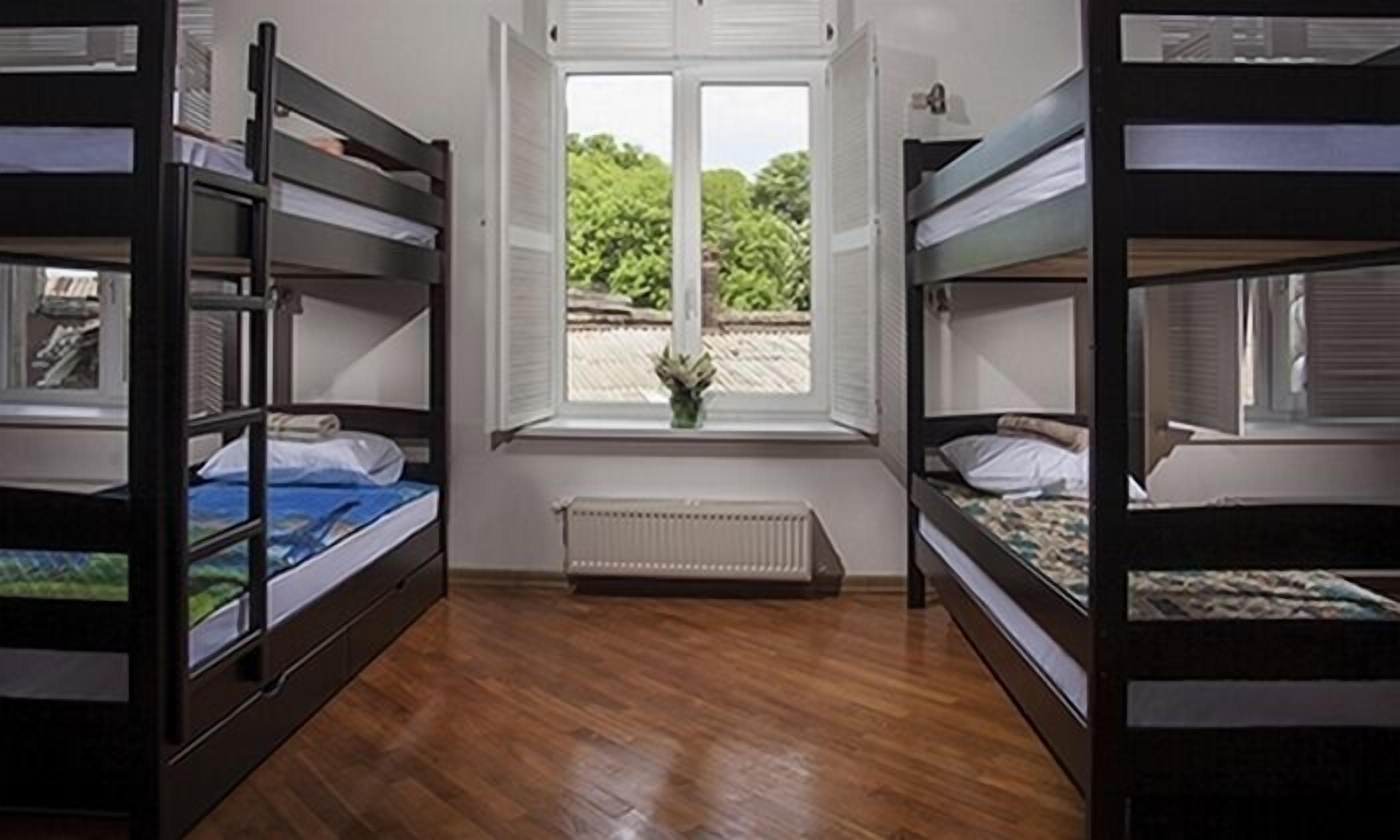 A dormitory room with bunk beds, a window with a view of greenery, and a radiator.