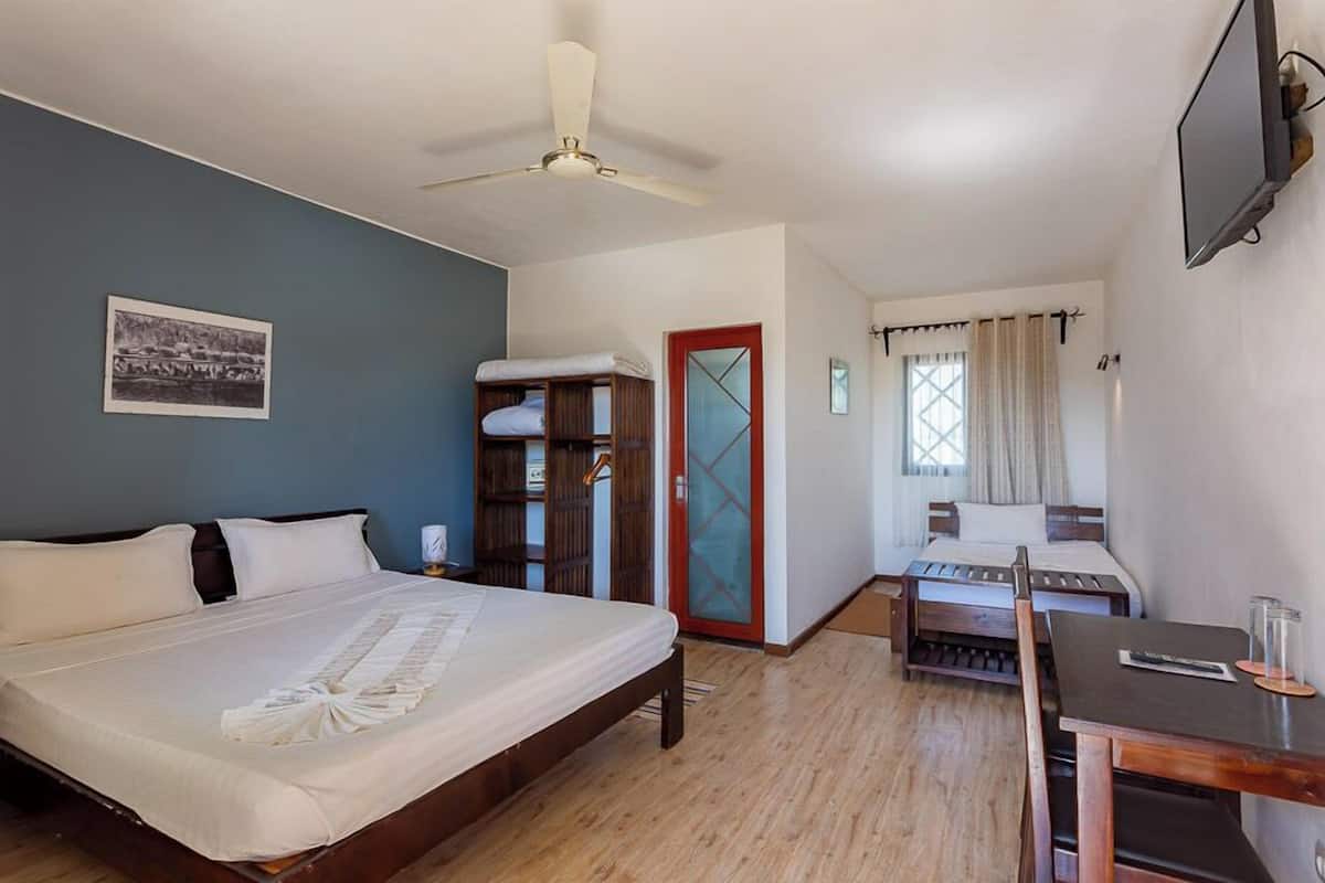 Une chambre d’hôtel avec un grand lit, un bureau, une chaise, un ventilateur de plafond, une télévision et une porte avec un panneau en verre.