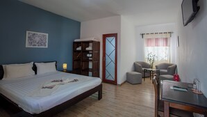 A hotel room with a bed, bedside table, a small seating area with a table and chairs, a bookshelf, and a window with curtains. - Hotel H1 Manakara SCHM (Manakara)