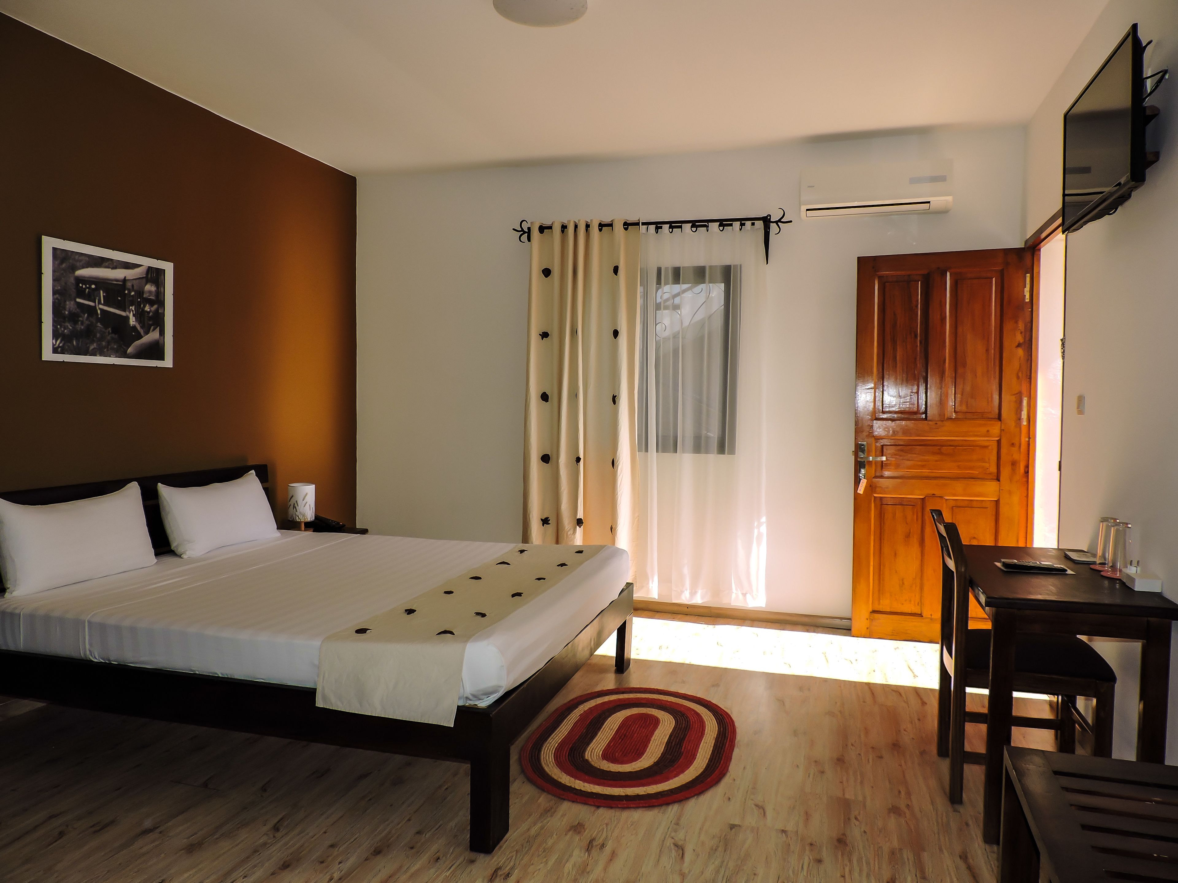 Un dormitorio con cama, puerta de madera, escritorio con silla y ventana con cortinas.